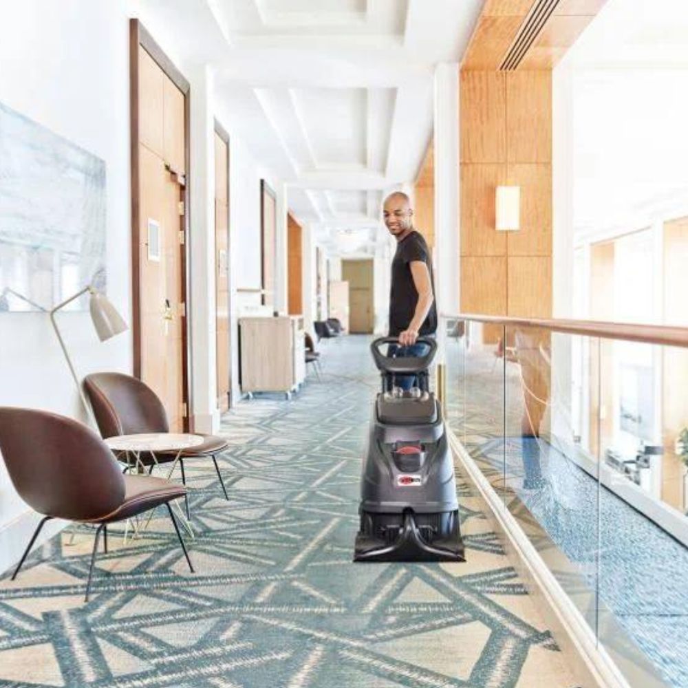 Man operating a floor cleaning machine on a patterned carpeted hallway.
