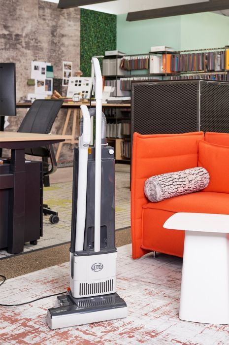 Upright vacuum cleaner on a patterned rug in an office setting with orange sofa and desk.