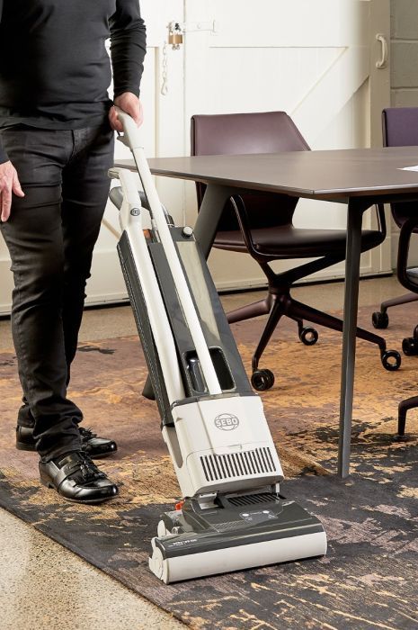Person vacuuming a patterned rug in an office with a table and chairs.