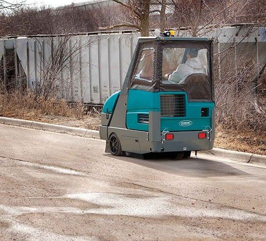 Teal and gray industrial sweeper with side brushes, driver seat, and headlights. 