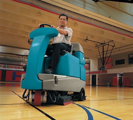 Turquoise and gray industrial floor scrubber with a seat and steering wheel.