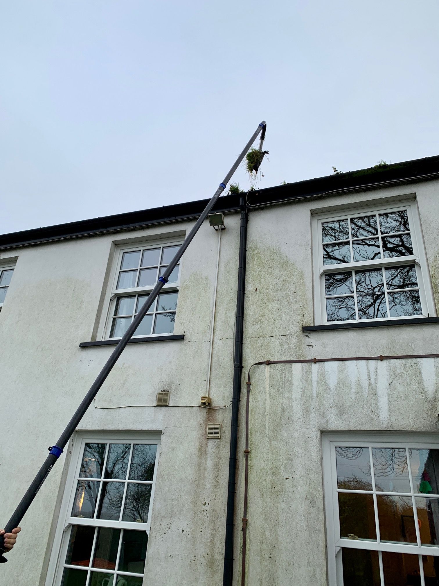 A person using a long-handled tool to clean the gutters of a two-story white building.