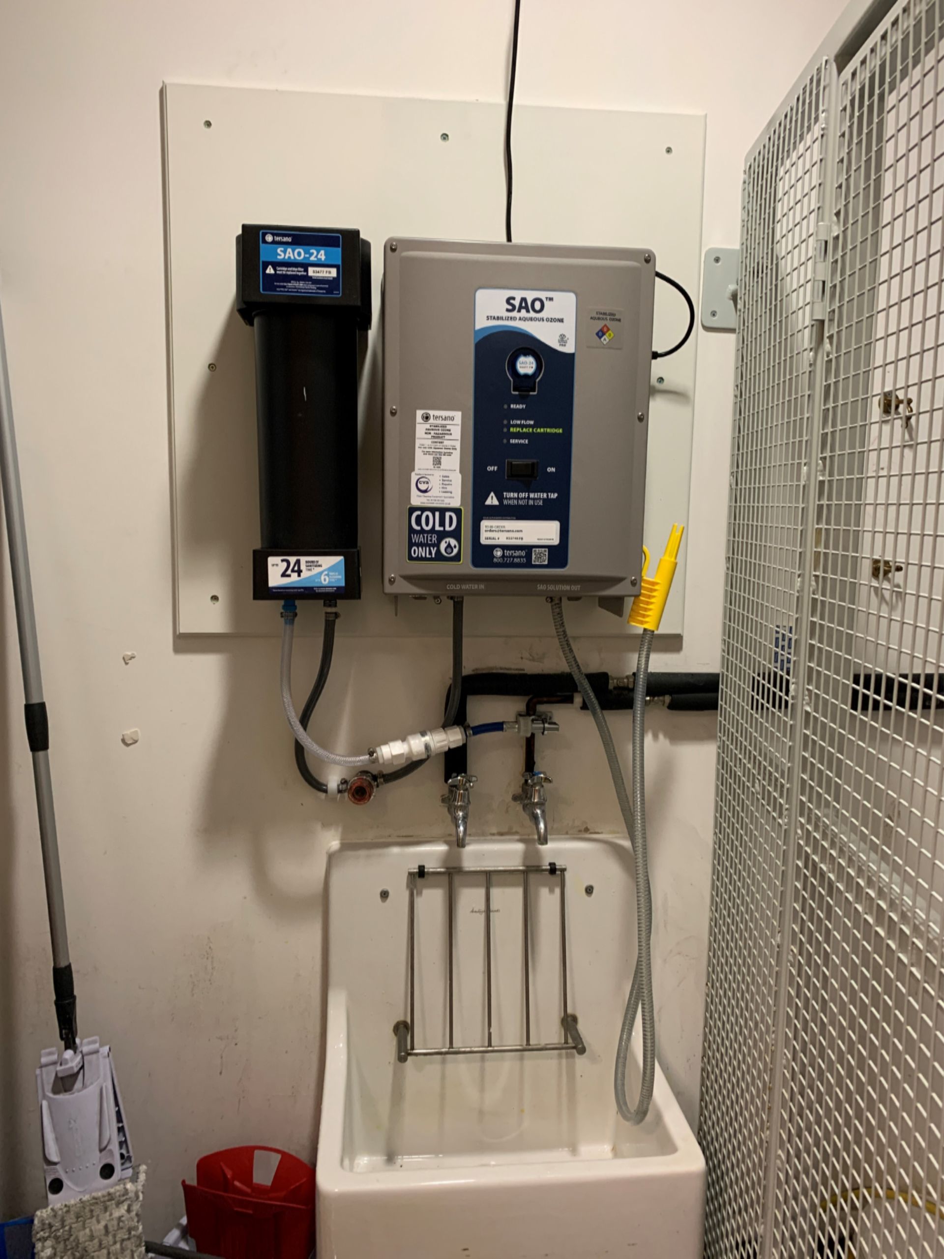 Water filtration and disinfection system mounted on a white panel above a utility sink. Black filter and gray control box.
