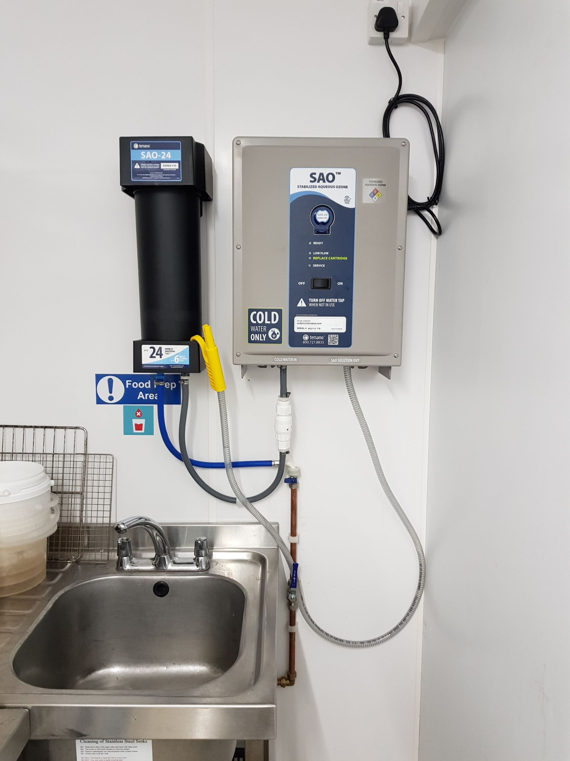Water filtration system above a stainless steel sink, mounted on a white wall.