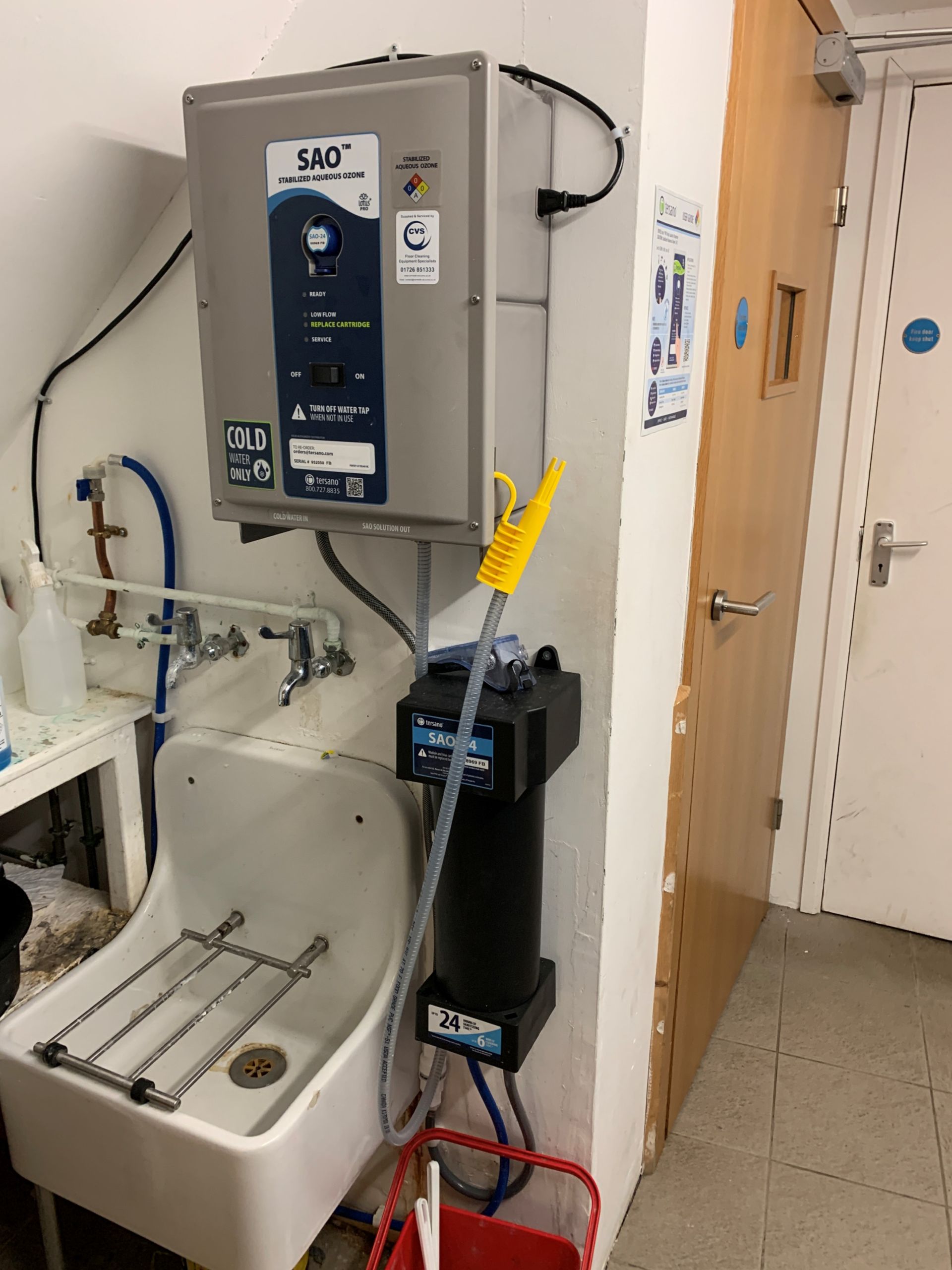 Sink with industrial equipment attached, including a water filter, against a wall next to a wooden door.