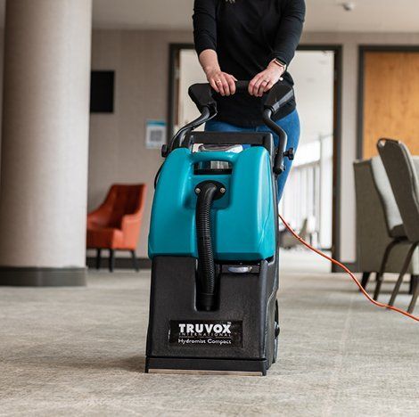 Woman operating a teal and black Truvox Hydromist Compact carpet cleaner in a hallway.