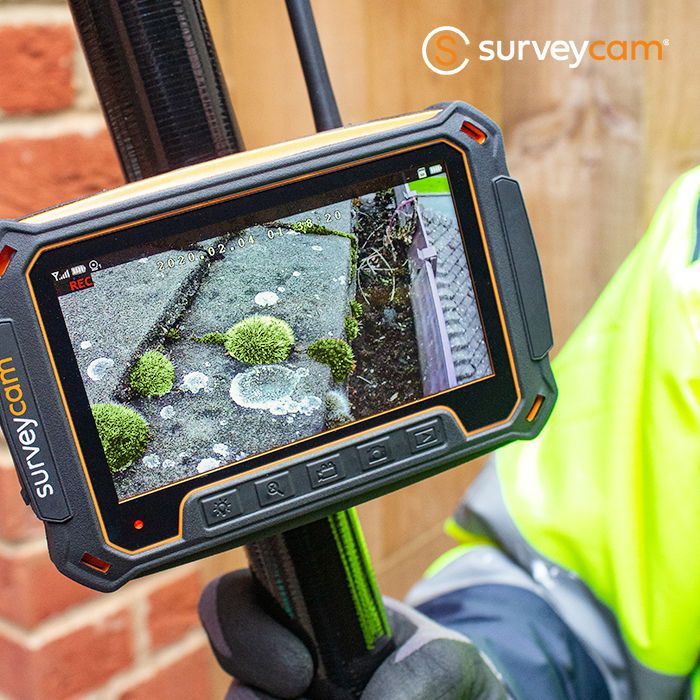 Handheld survey cam displaying a close-up of a mossy surface. The operator wears work gear and holds the device near brick.
