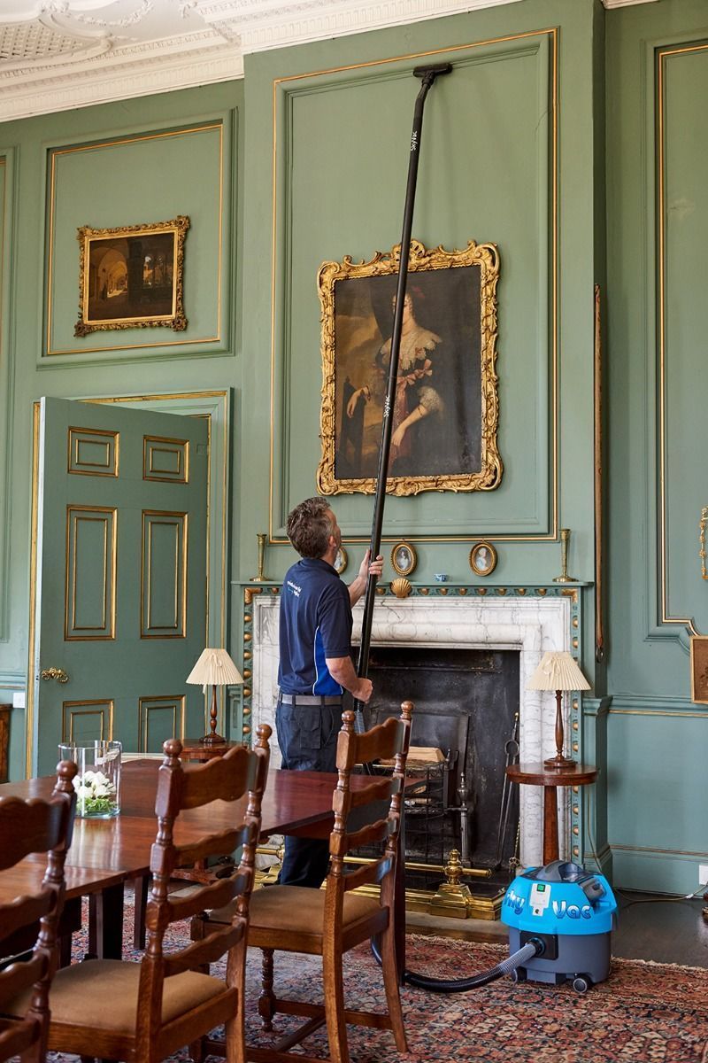 Man vacuuming a wall in a room with paintings and a fireplace. Blue vacuum cleaner on the floor.
