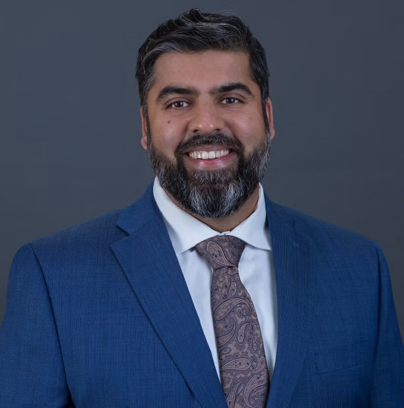 Man in blue suit, smiling, with a grey beard and patterned tie against a grey background.