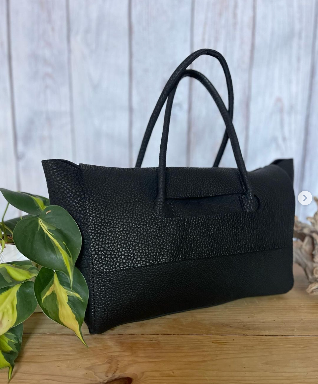 A black purse is sitting on a wooden table next to a plant.