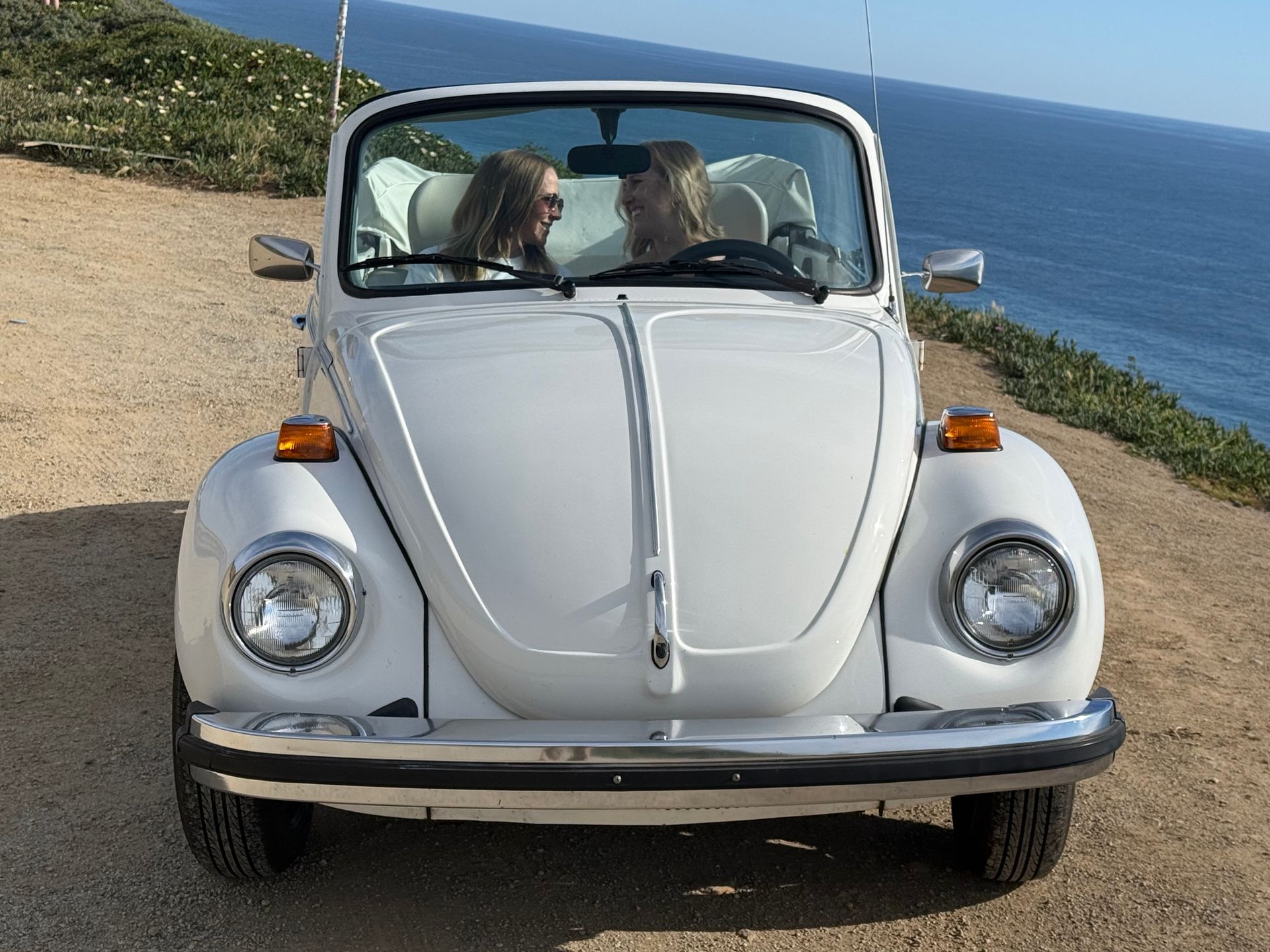 A couple in wedding attire poses in a red convertible by the ocean, with the bride’s feet resting on the dashboard.