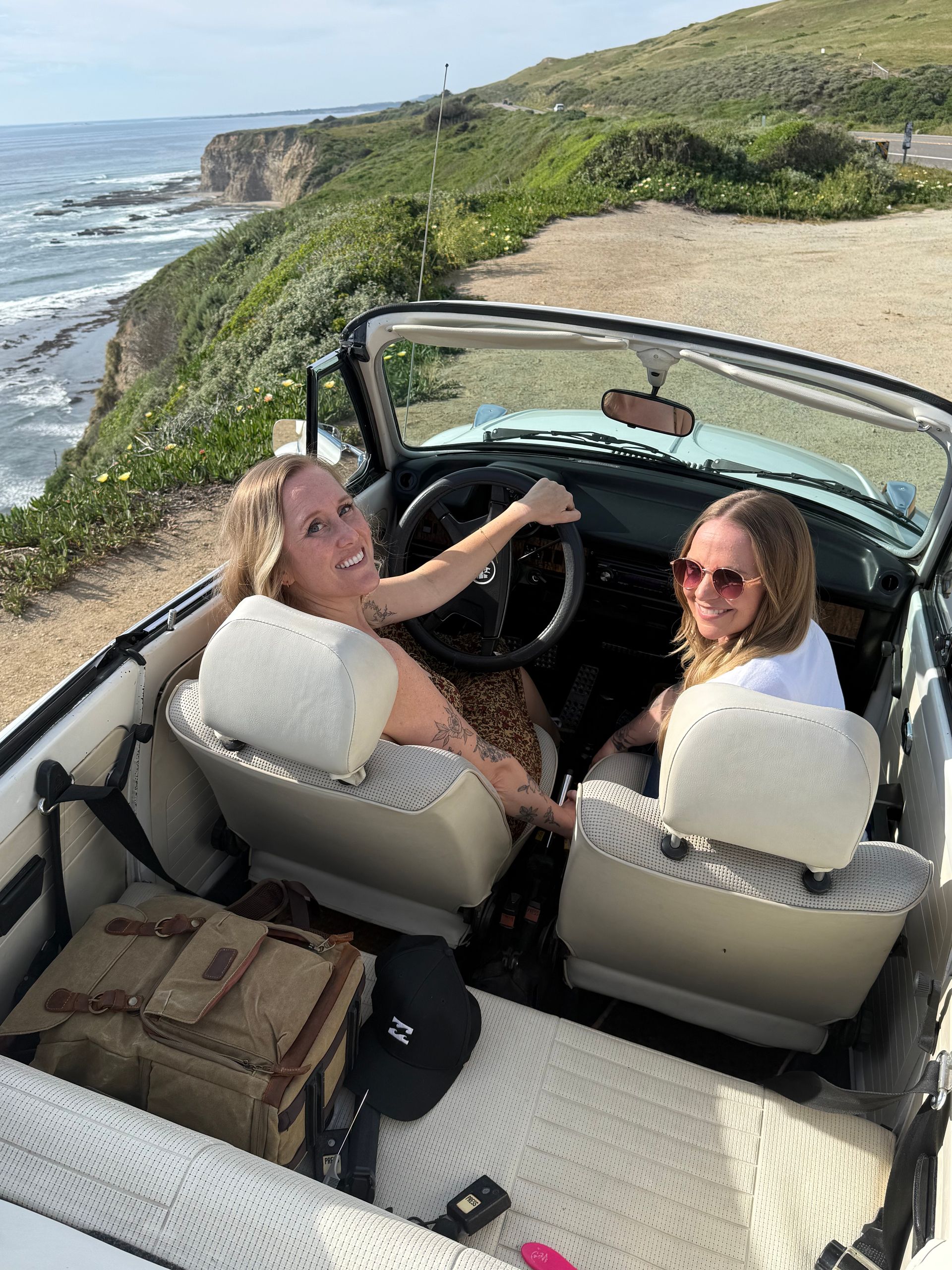 A couple stands by a red vintage convertible parked on a gravel lot overlooking the ocean at sunset.