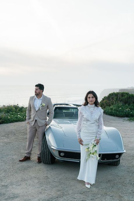 A person in a tan suit and a person in a white lace dress stand in front of a silver convertible overlooking the ocean.