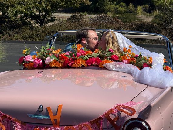 A newlywed couple kisses in a pink convertible decorated with bright flowers and a 