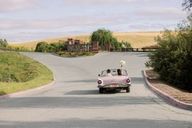 A couple in a vintage pink convertible drives away on a road in a scenic, hilly landscape, one waving a bouquet.