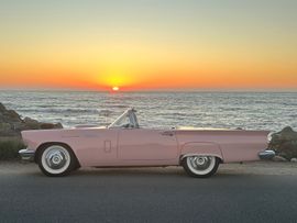 A pink vintage convertible parked on the side of a road overlooking the ocean at sunset.