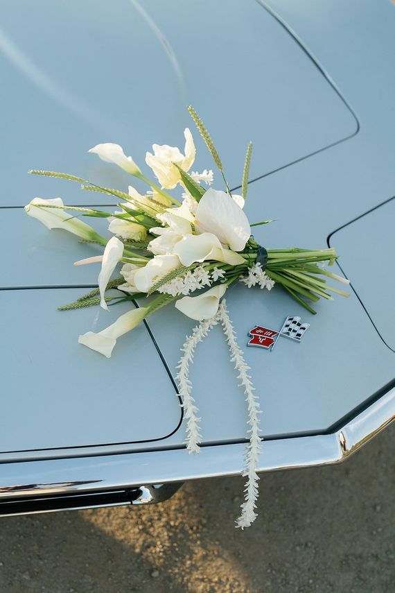 A bridal bouquet of white calla lilies and greenery rests on the light blue hood of a vintage car with two small emblems.