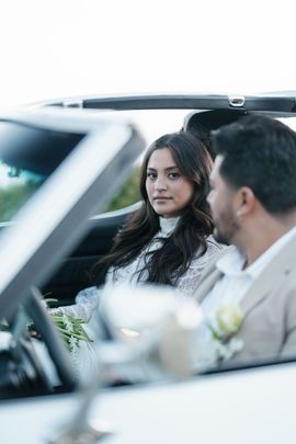 A couple in wedding attire sitting in a convertible, with the woman looking forward and the man looking at her.