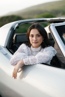 A person with dark hair wearing a white lace top, leaning against the side of a silver convertible outdoors.