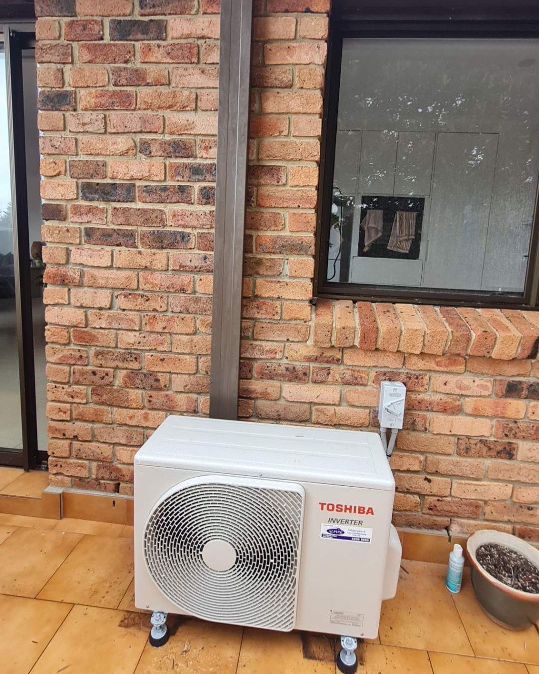 Outdoor Air Conditioner Unit on Tiled Patio Beside a Brick Wall and Window — Alpine Refrigeration & Air Conditioning Pty Ltd in Wollongong, NSW