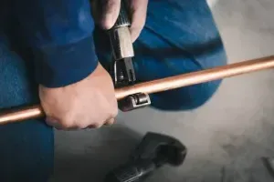 Hands Using A Pipe Cutter On A Copper Pipe Over A Concrete Floor — Alpine Refrigeration & Air Conditioning in Wollongong, NSW Hands Using A Pipe Cutter On A Copper Pipe Over A Concrete Floor — Alpine Refrigeration & Air Conditioning in Wollongong, NSW