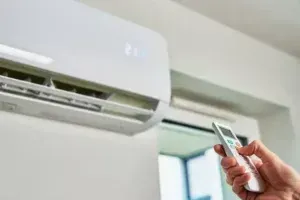 Hand Holding A Remote Pointed At A Wall Mounted Air Conditioner In A Bright Room — Alpine Refrigeration & Air Conditioning in Wollongong, NSW