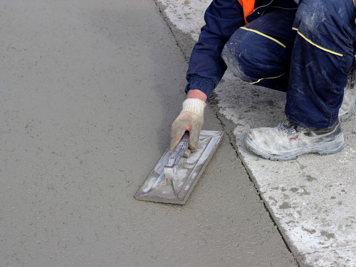 A construction worker in blue overalls using a trowel to smooth wet concrete.