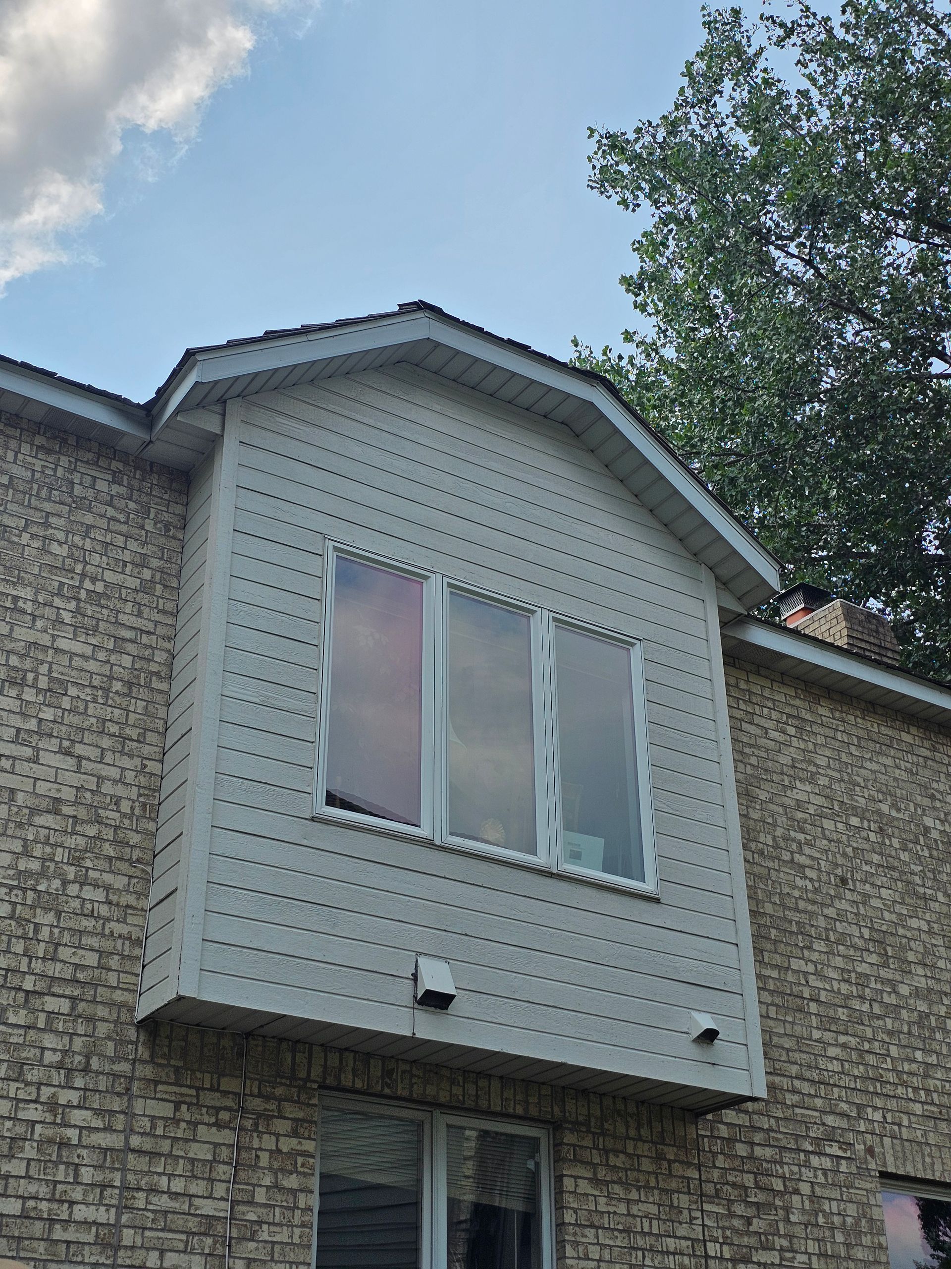Bay window with three panes on a brick building, under a light blue sky and a tree.