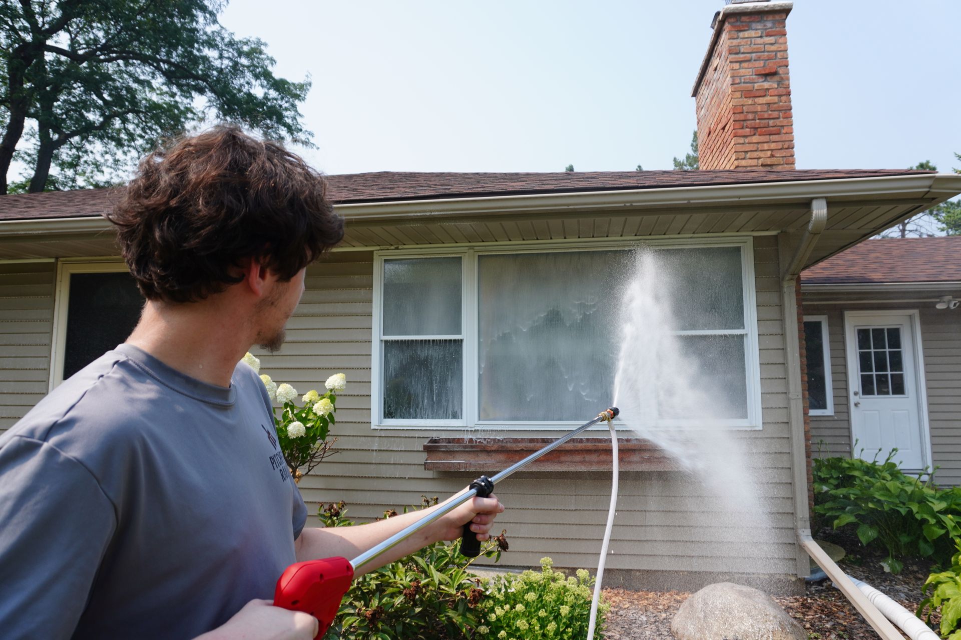 Man spraying water at a window of a beige house with red brick chimney and green bushes.