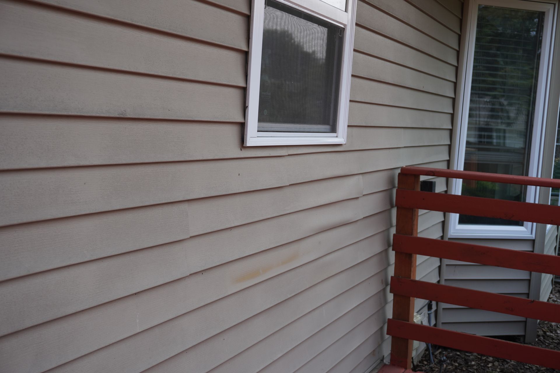 Beige siding of a building with a white-framed window. A red railing is visible on the right.