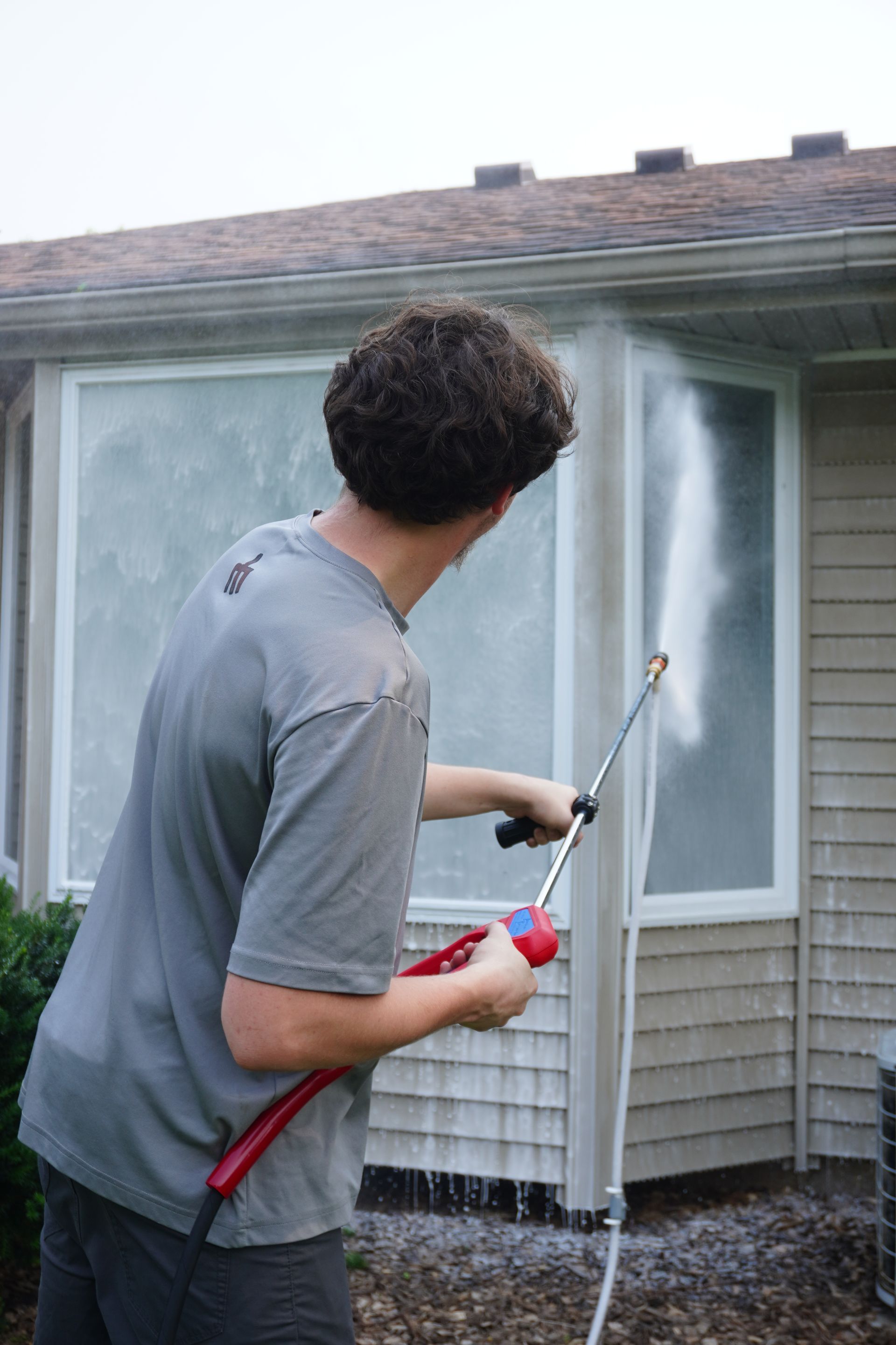 Man pressure washing a house exterior near a window.