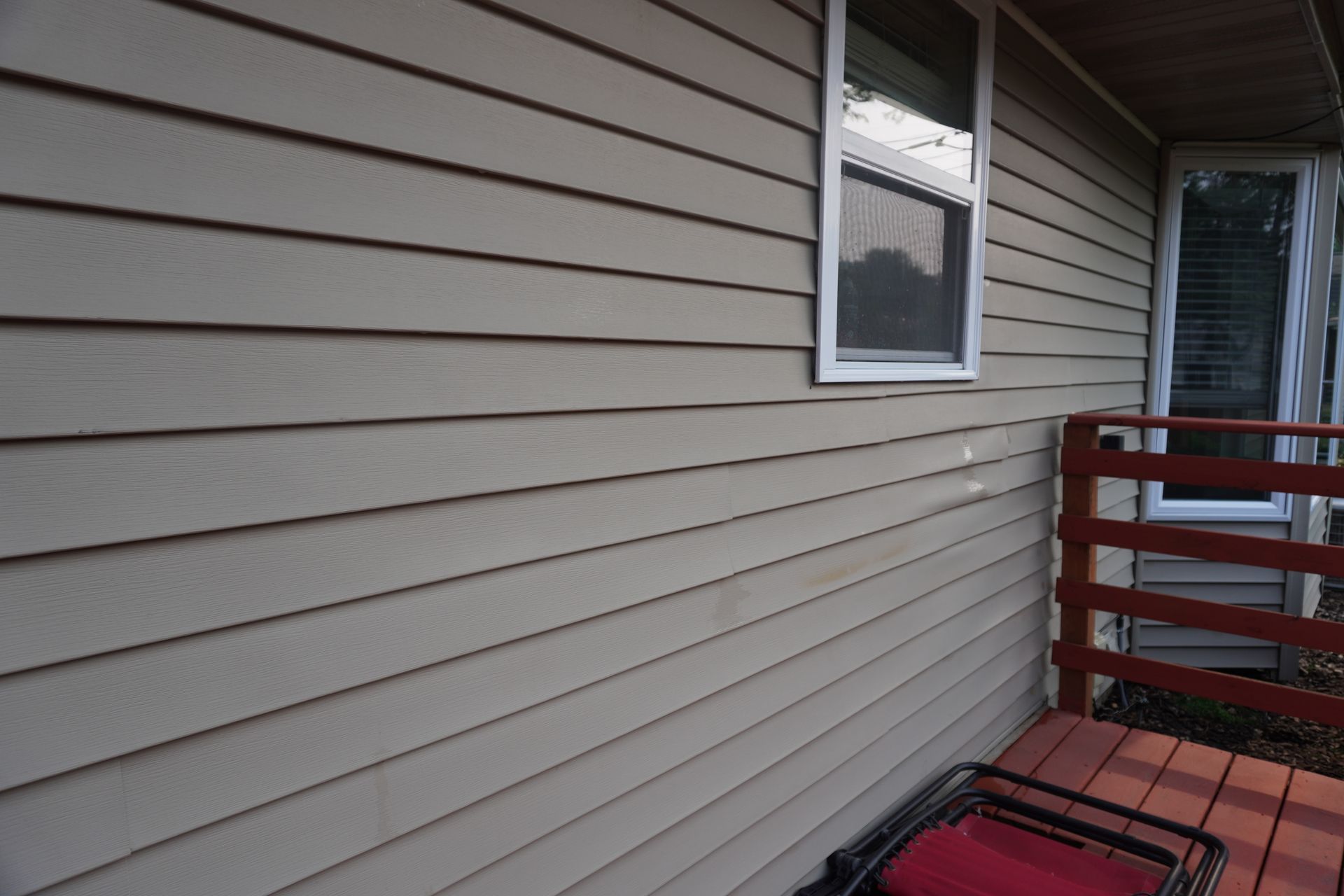 Beige siding on a house with windows and a red deck railing. A chair sits on the deck.
