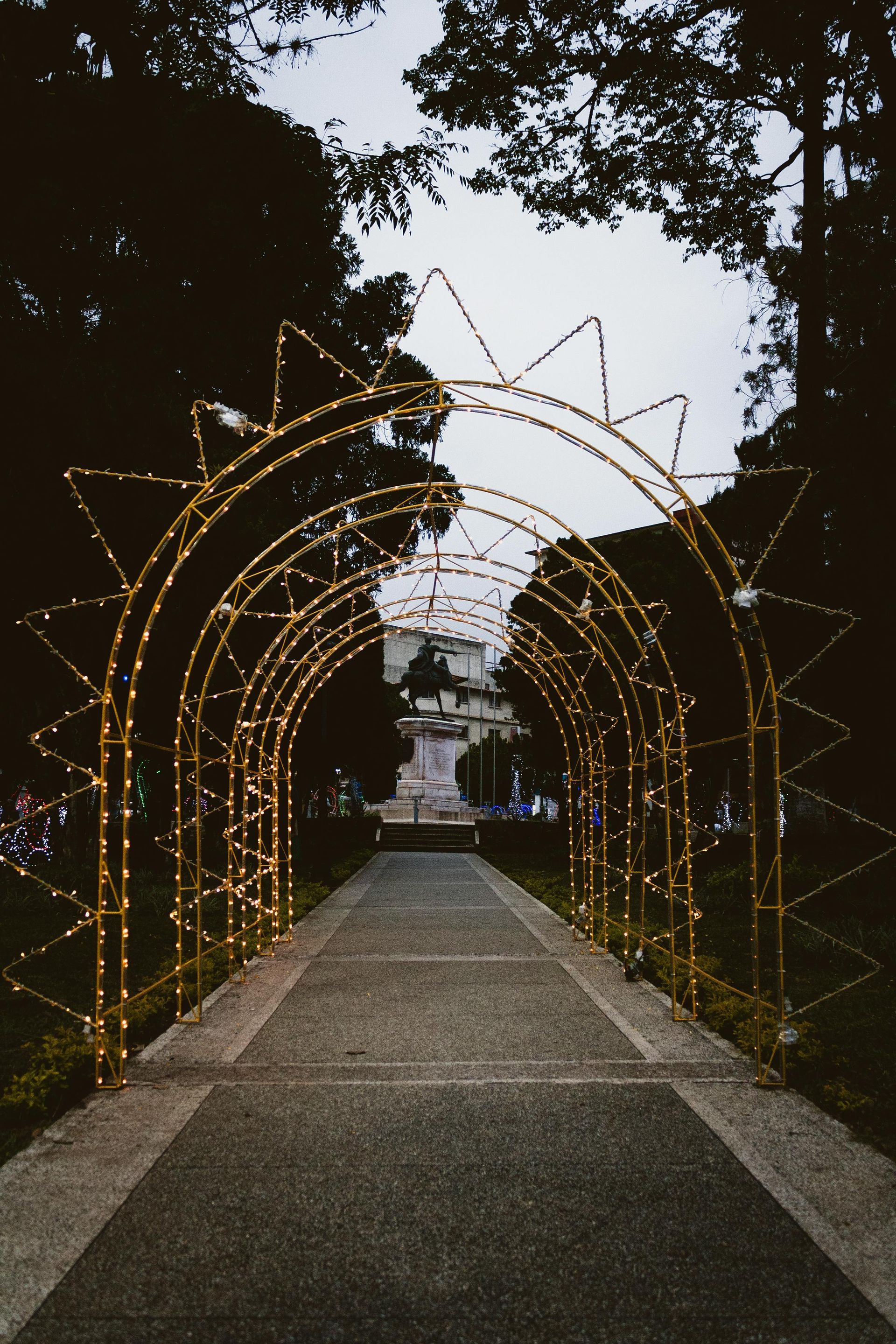 Path through arches with lights, leading to a statue in a park.