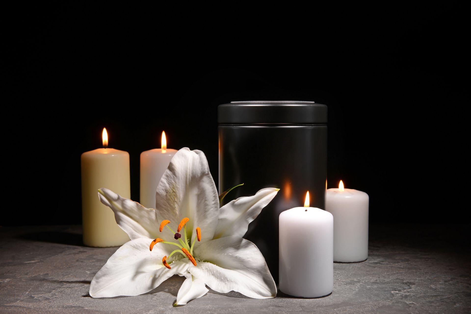 Black memorial urn with white lily and lit candles arranged on a dark surface for cremation remembrance.