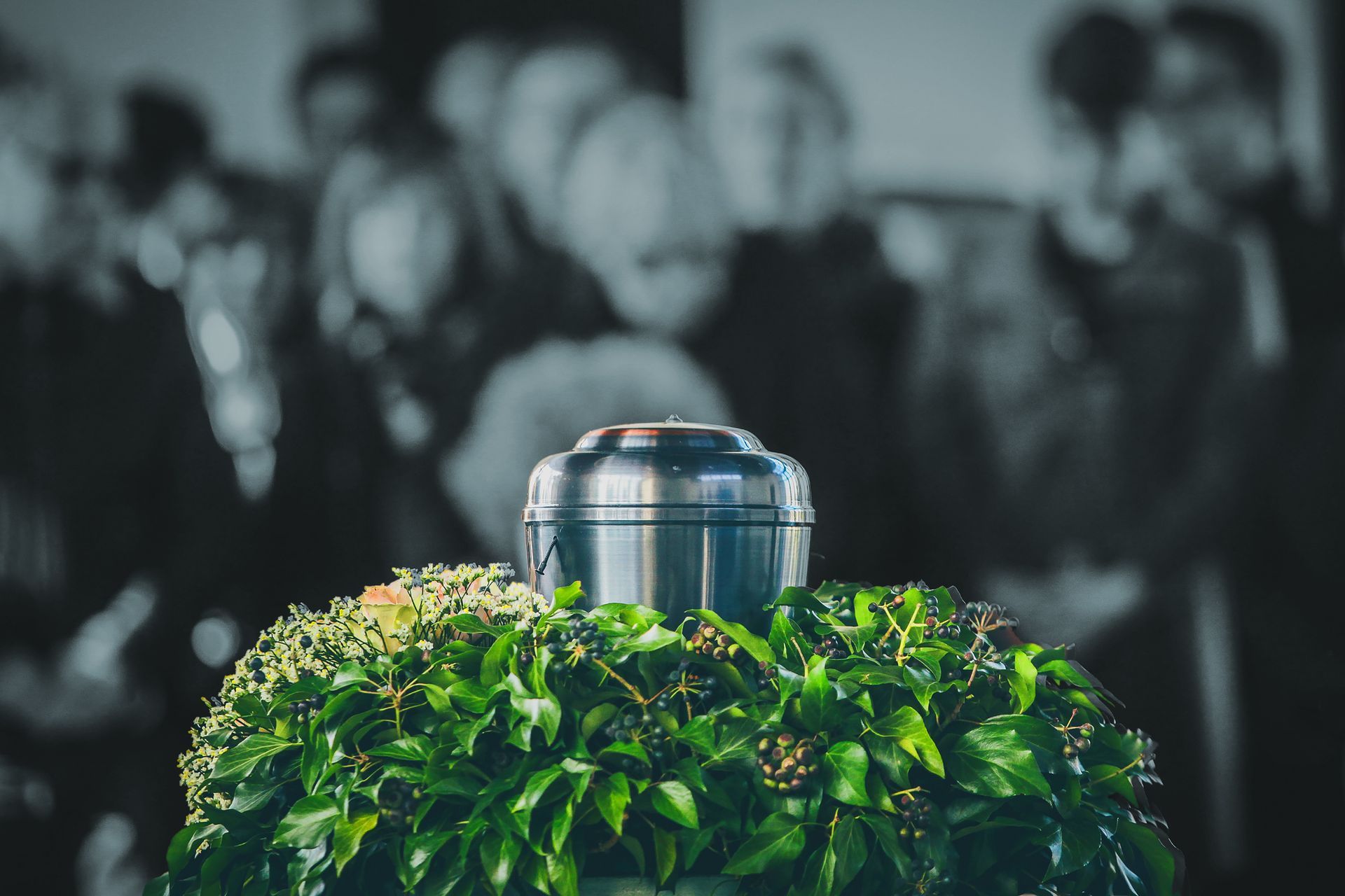 Silver urn resting on green foliage at a memorial, with blurred attendees in the background.