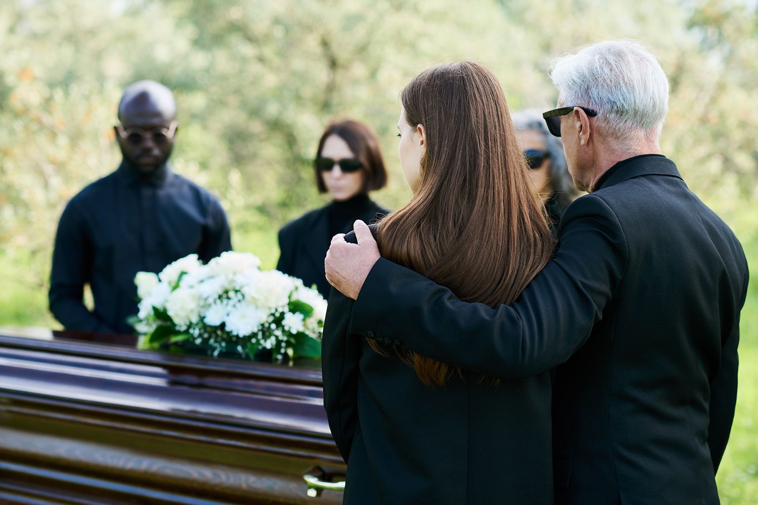 People gathered at a funeral beside a casket adorned with white flowers.