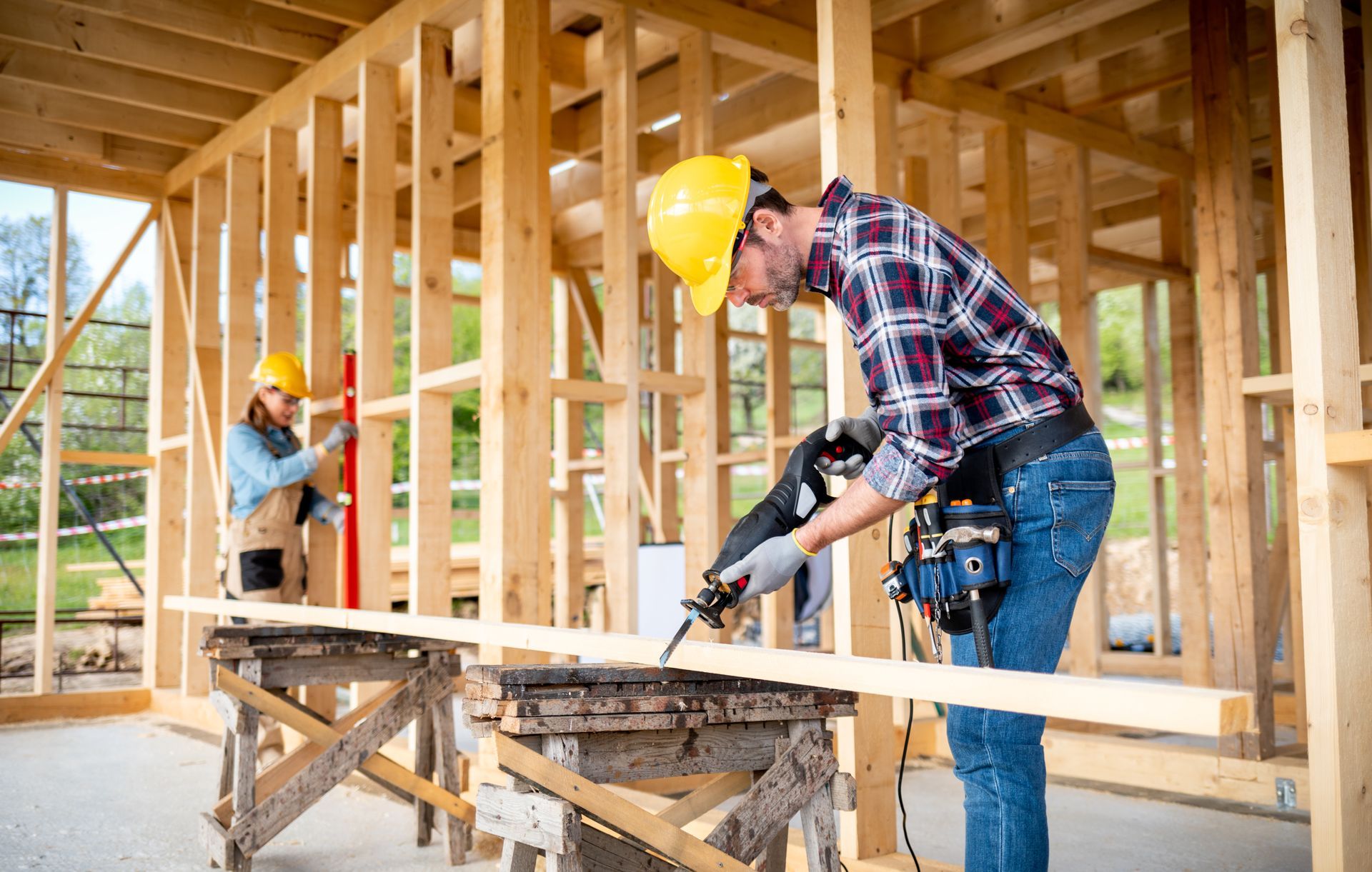 Two construction workers at a building site; one cutting wood, the other using a level.