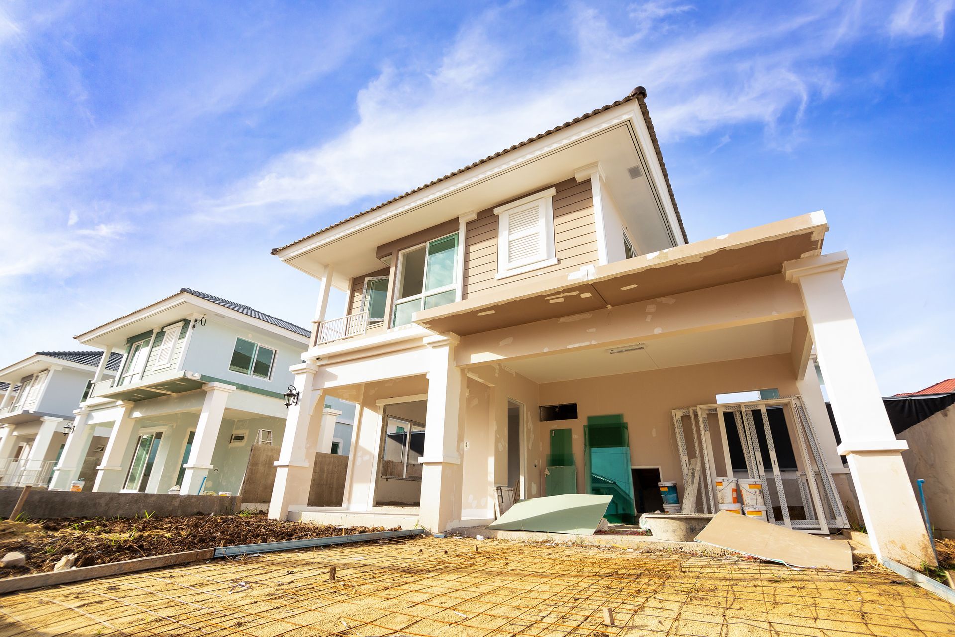 Two-story beige house with a covered porch and construction materials in the front yard; blue sky.