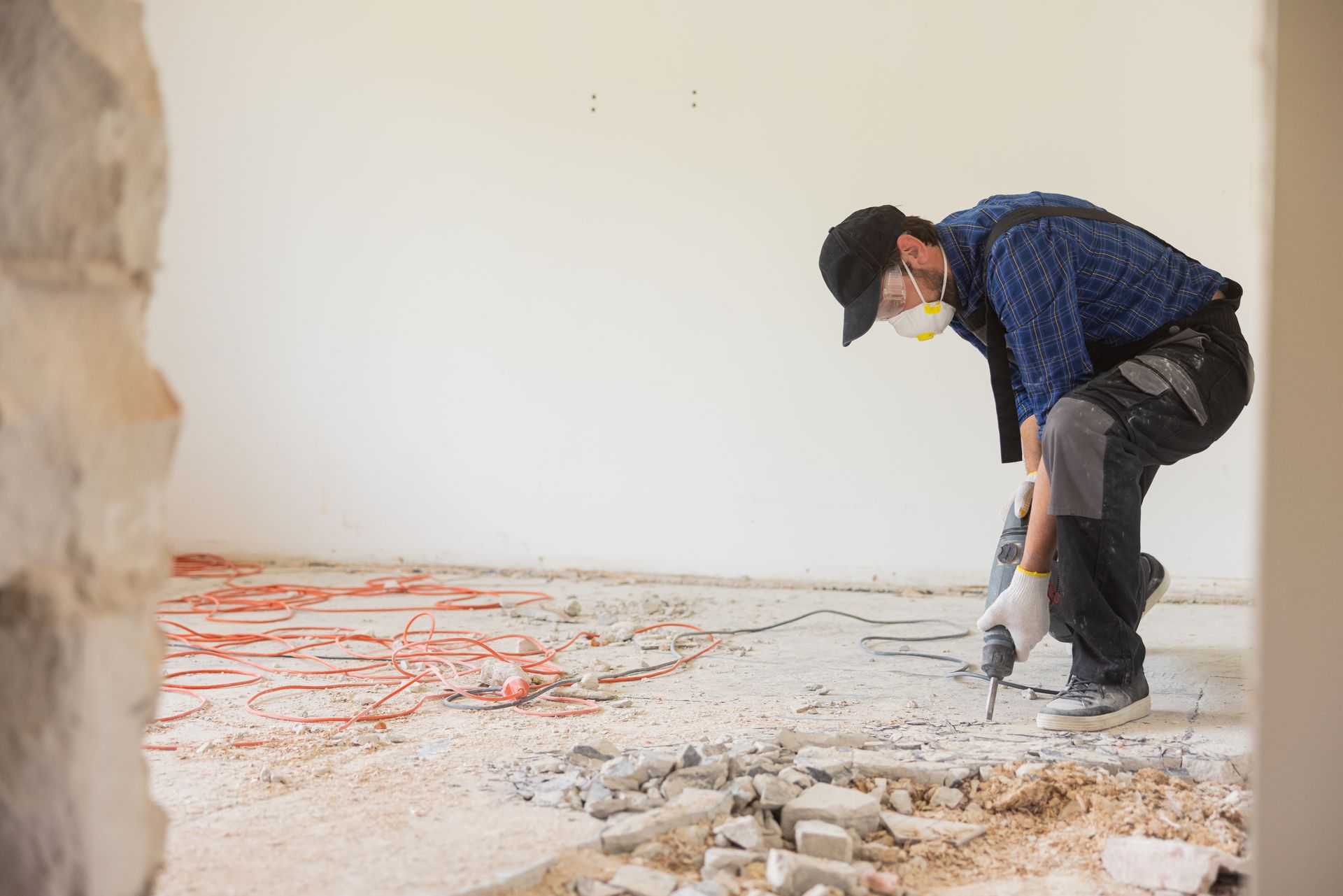 Person in safety gear using a jackhammer to break up a concrete floor in a room.
