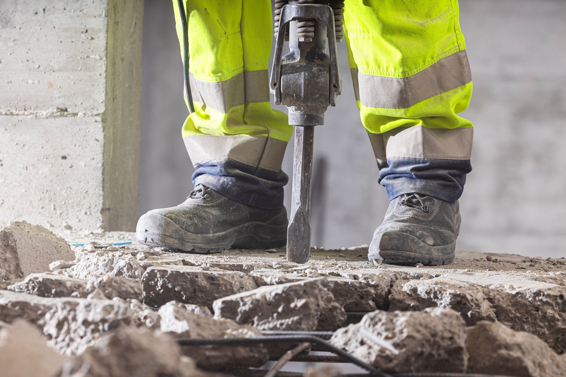 Construction worker using a jackhammer on concrete; wearing yellow safety gear and work boots.