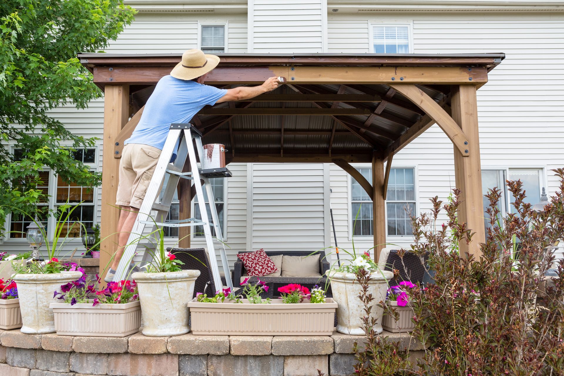 Man on ladder paints wooden gazebo near a house with flower boxes.