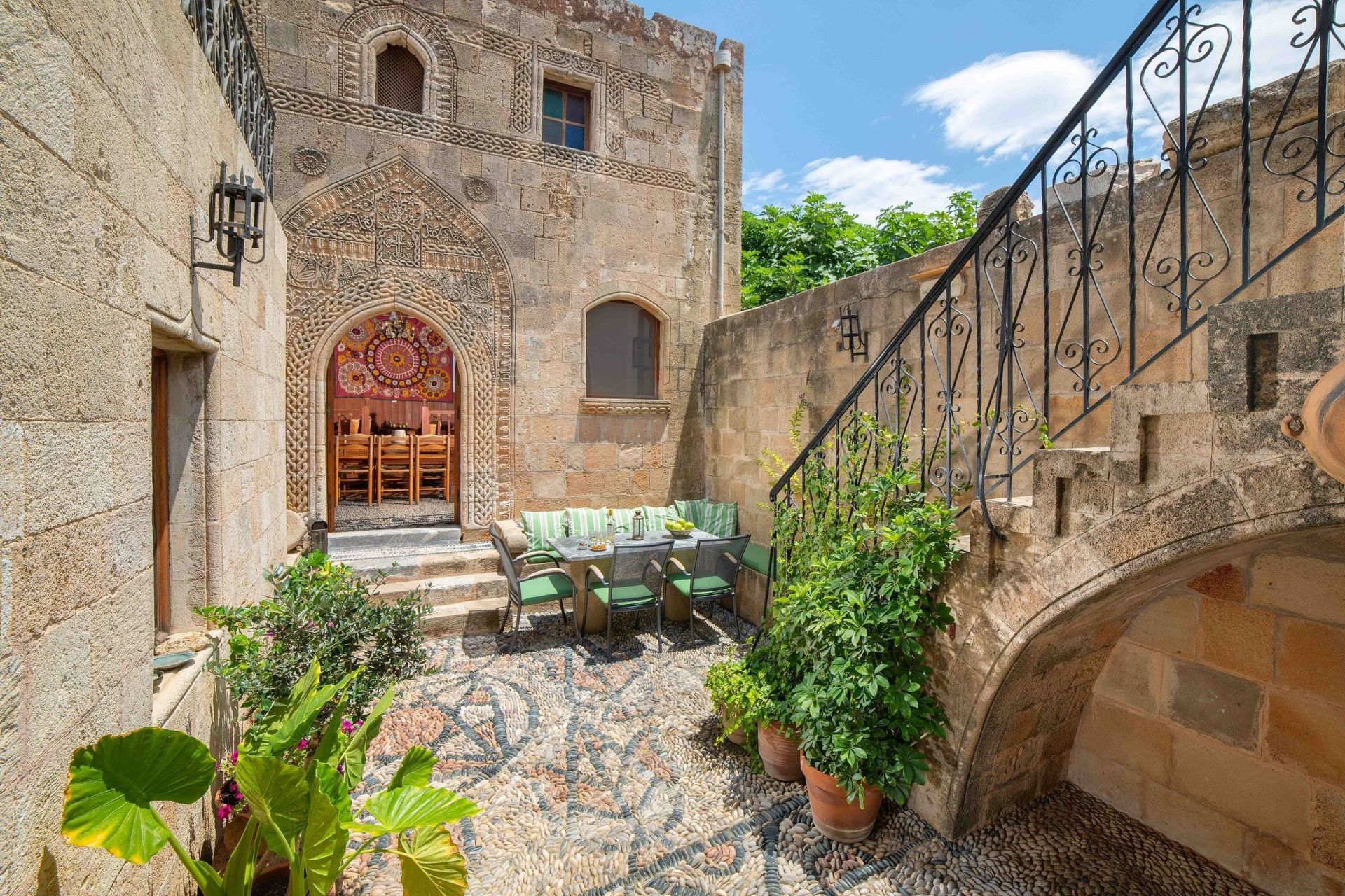 Stone courtyard with arched stairway, wooden door, and potted plants in bright sunlight