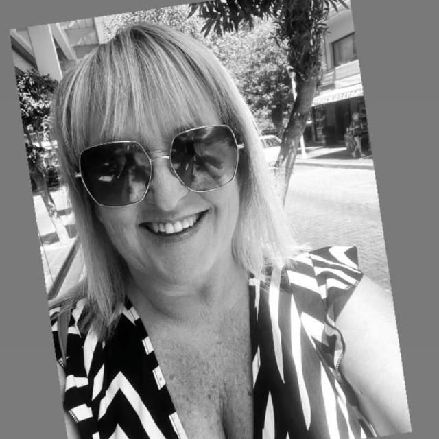 Smiling person wearing sunglasses and a patterned top, taking a selfie outdoors.