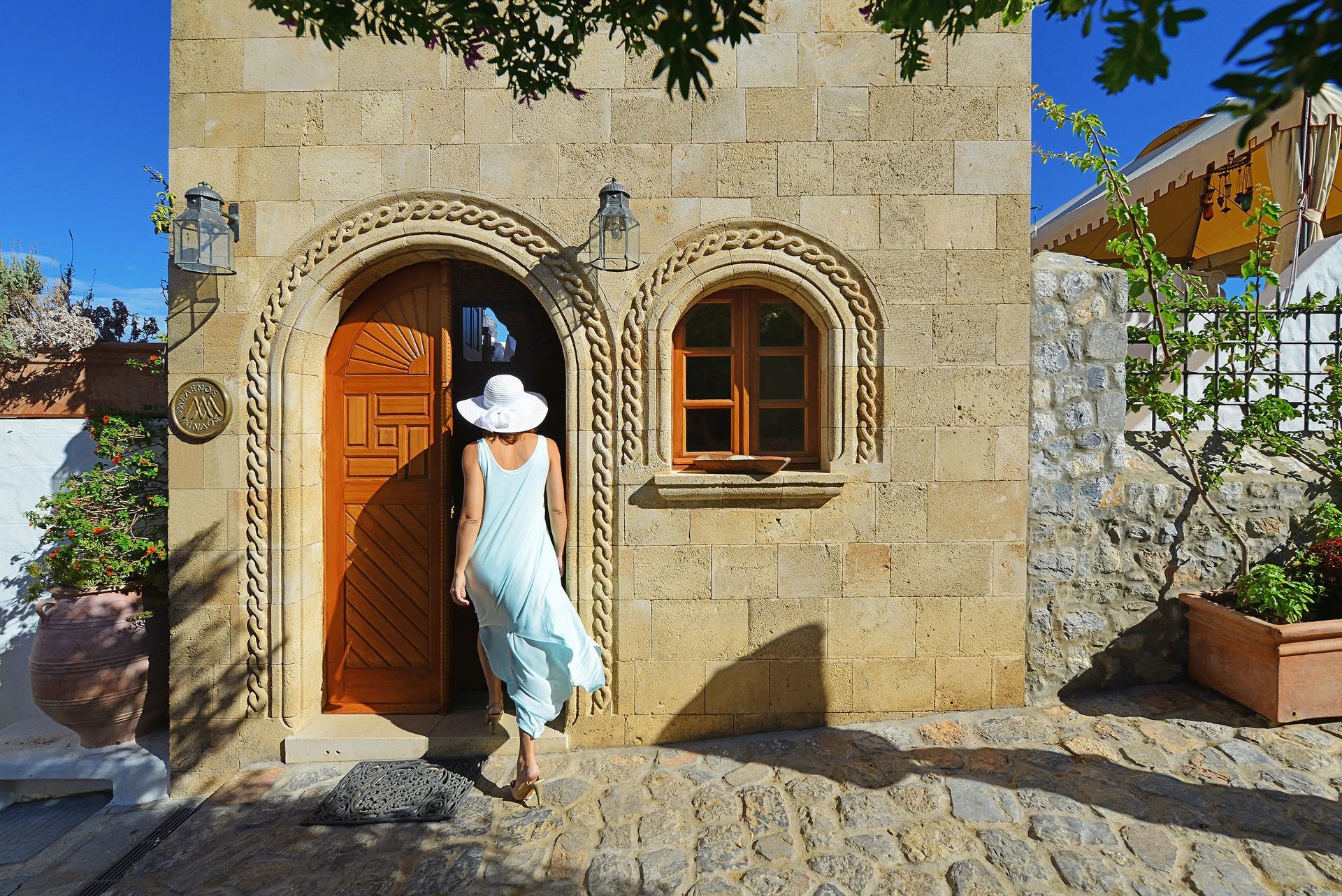 Person in a white hat and blue dress walks toward a rustic stone doorway with arched windows.