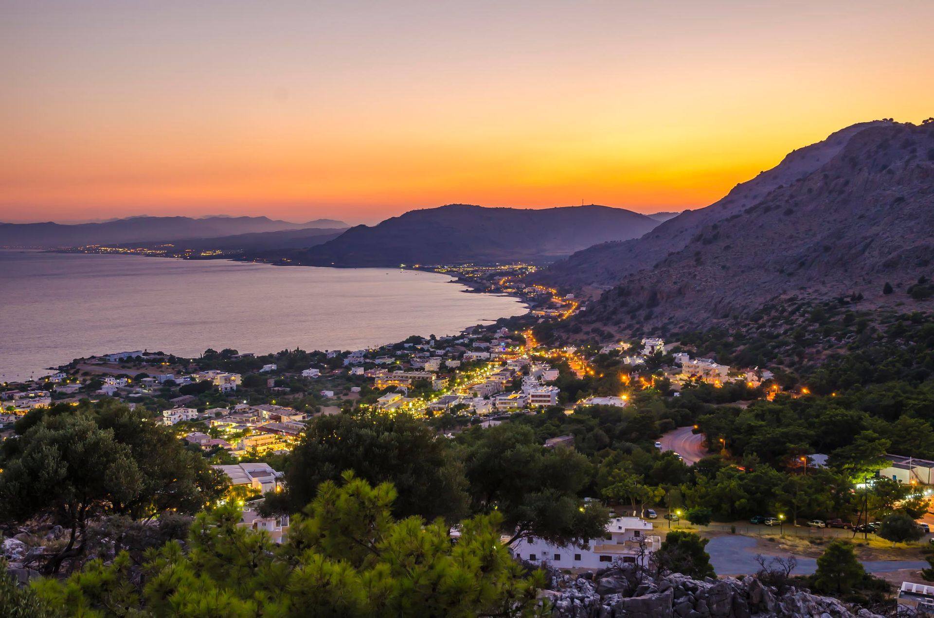 Sunset over a coastal bay with hills, houses, and city lights below a purple-orange sky