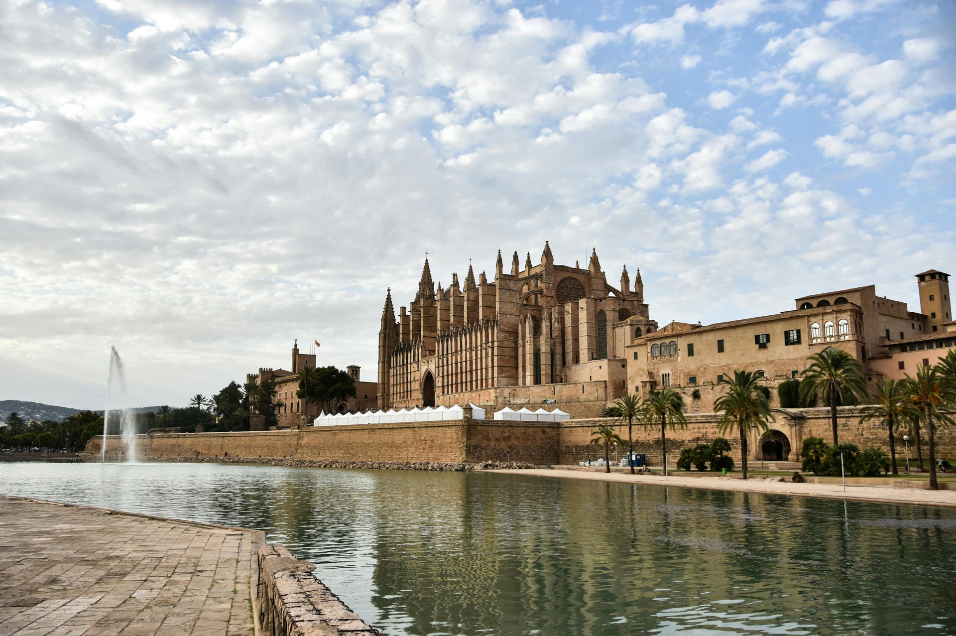 Gothic cathedral beside a reflective canal under a cloudy sky, with fountains in the foreground.