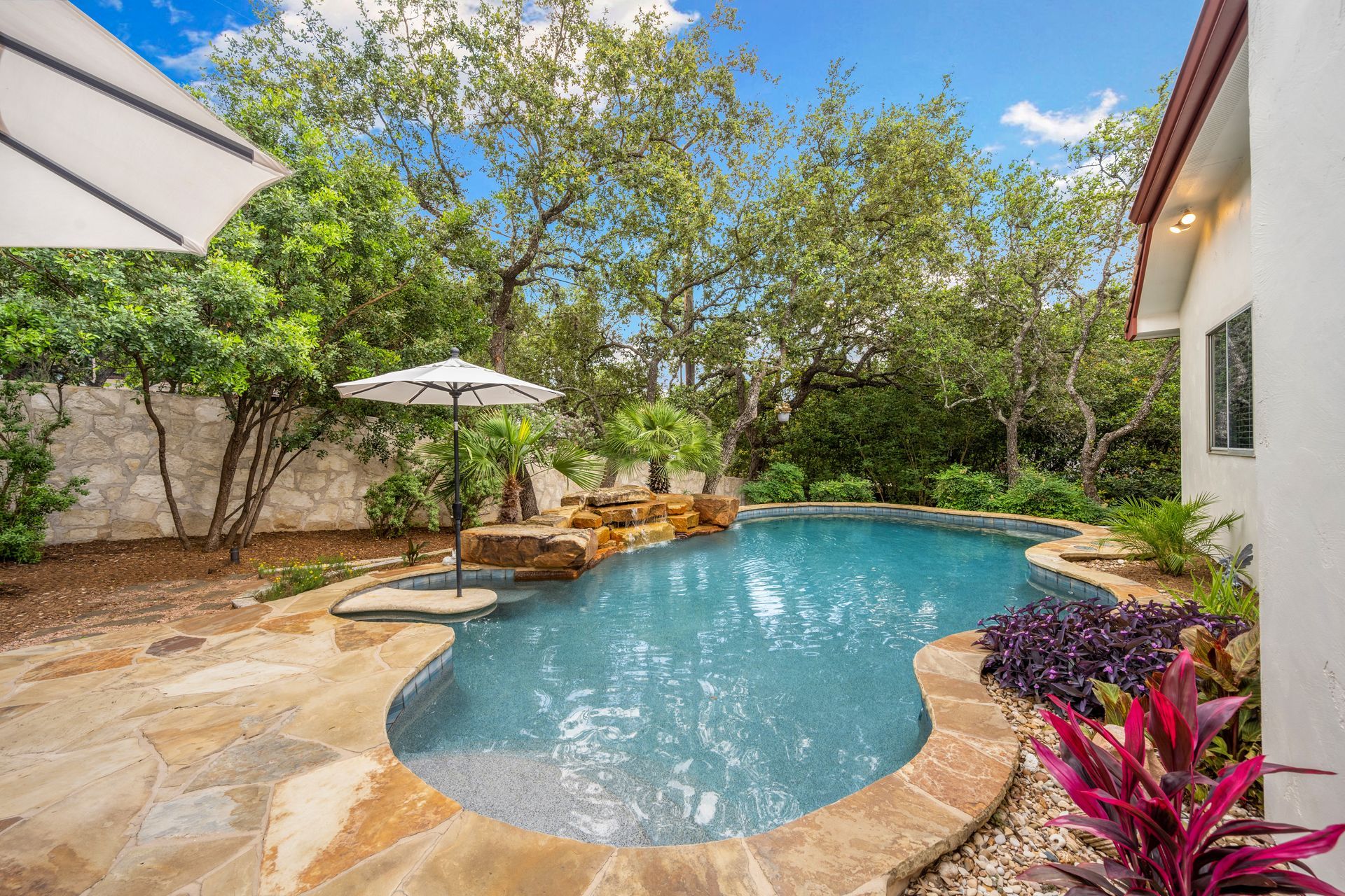 Backyard pool with stone patio, lounge chairs, umbrellas, and lush trees under a blue sky
