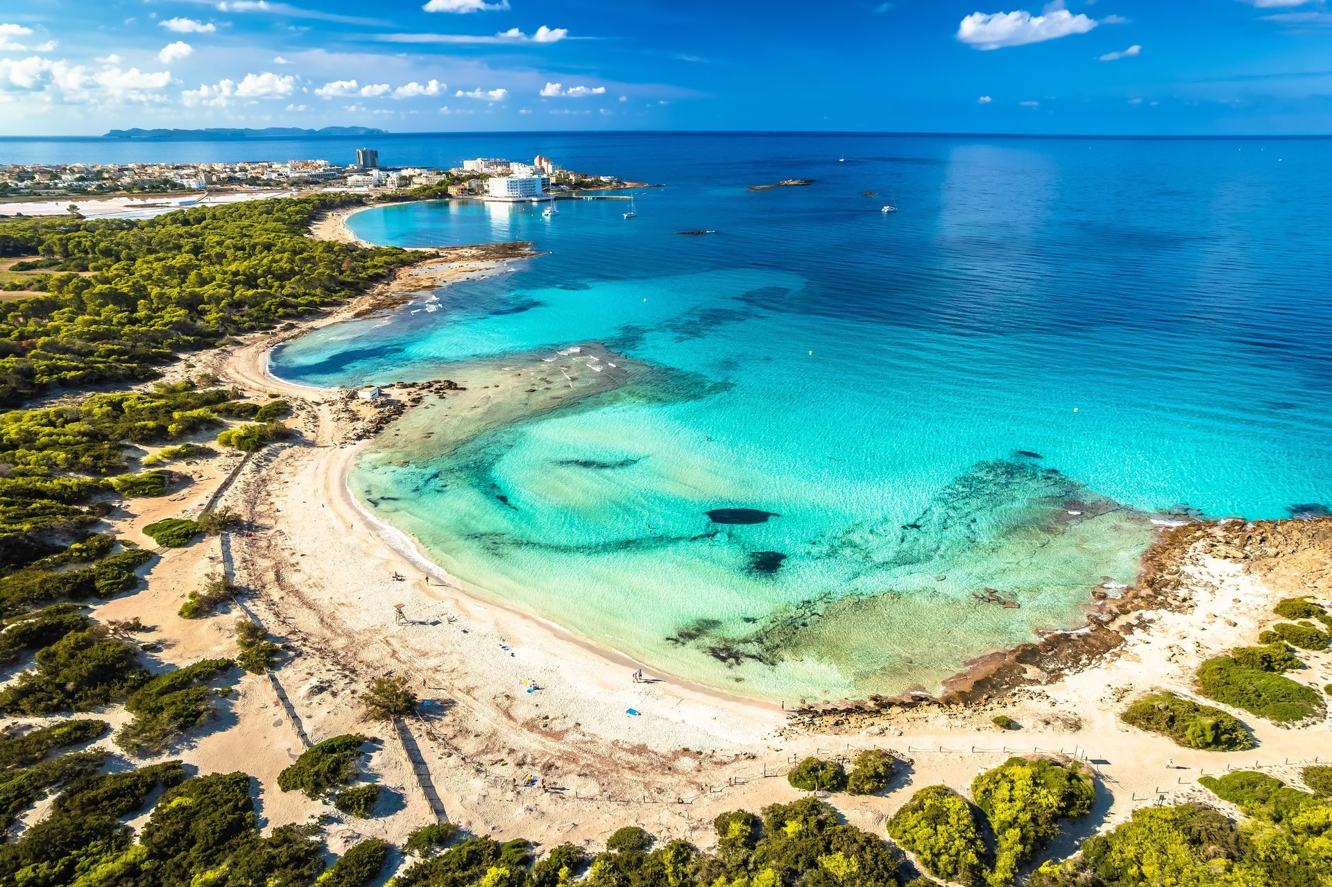 Aerial view of a turquoise tropical bay with sandy beach, rocky shoreline, and clear blue ocean under a sunny sky