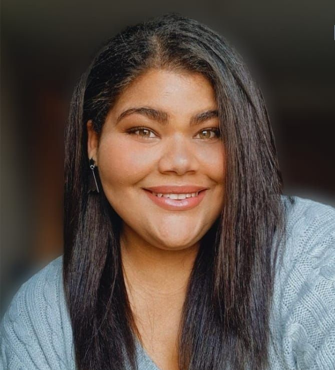 A woman with long black hair is smiling and wearing a blue sweater.