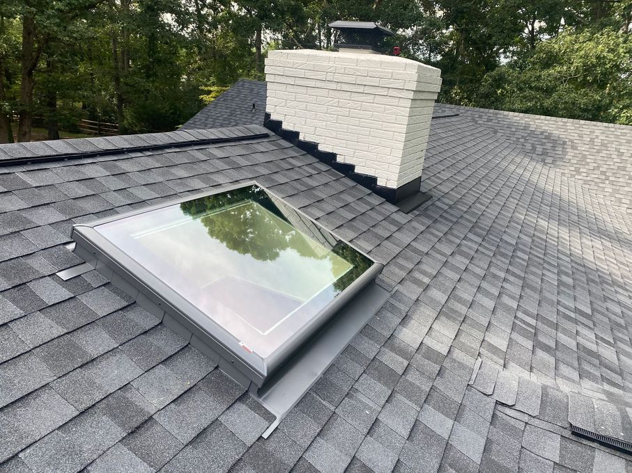 A modern skylight installed on a dark gray shingled roof next to a white brick chimney.